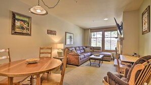 A cozy living room with a wooden dining table, a leather sofa, and a wooden armchair. There is a large window with curtains, a wall-mounted lamp, and a framed picture on the wall.