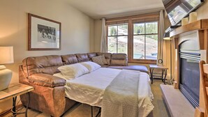 A cozy bedroom with a large bed, a leather sofa, a fireplace, and a large window with a view of a snowy landscape.