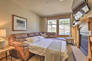 A cozy bedroom with a large bed, a leather sofa, a fireplace, and a large window with a view of a snowy landscape.