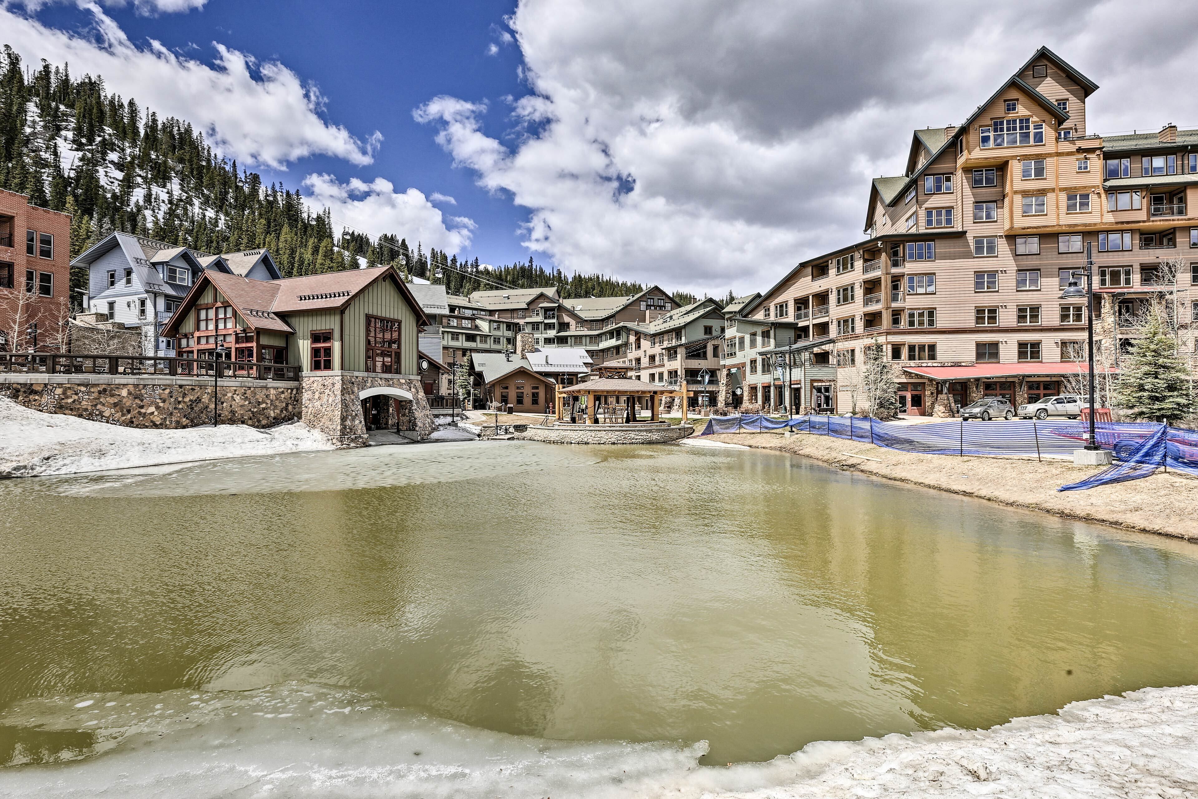A riverside hotel with a stone bridge, wooden buildings, and a backdrop of trees and mountains.