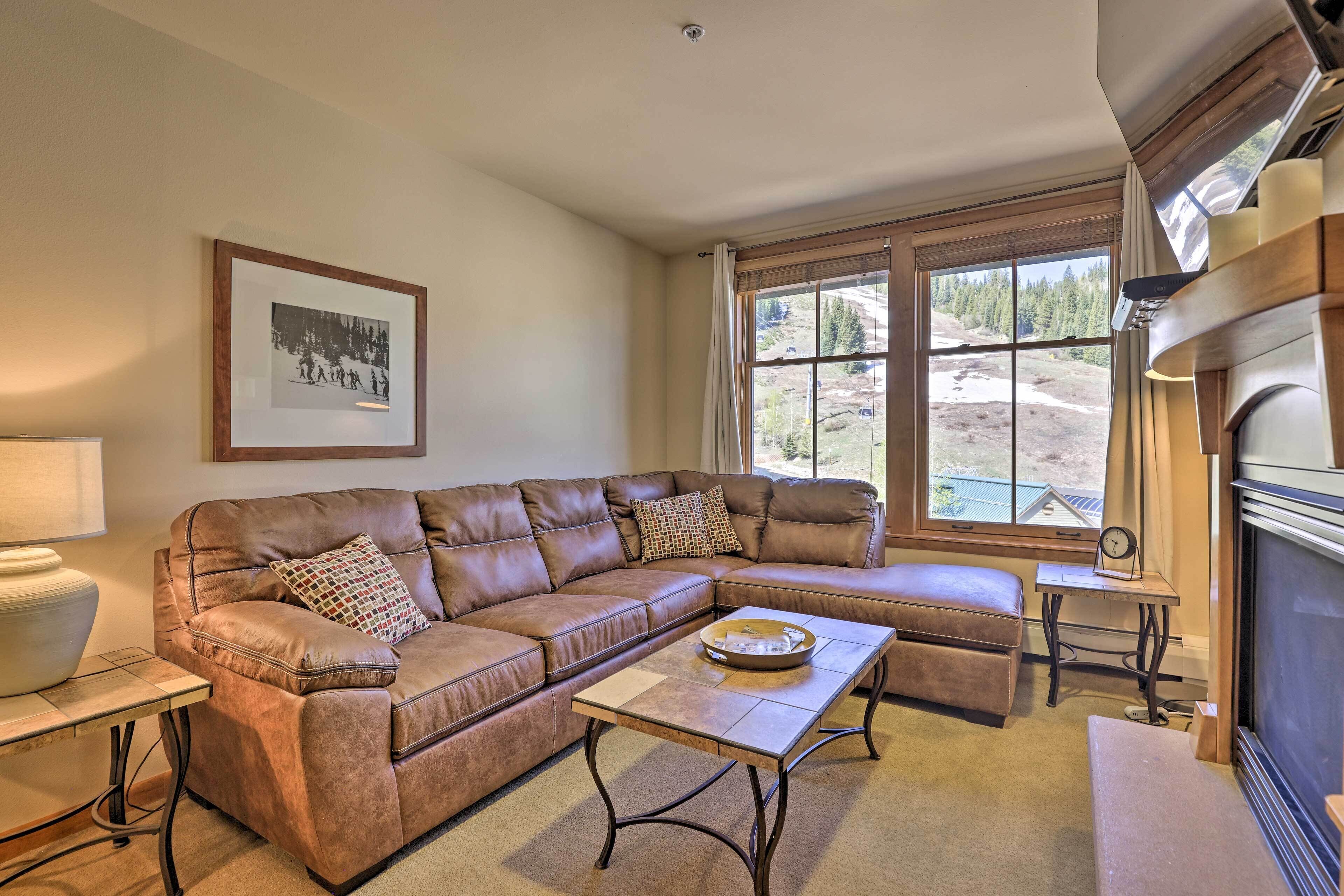 A living room with a brown leather sectional sofa, a coffee table, a fireplace, and a large window with a view of trees and a building.