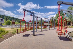 An outdoor area with swings and benches, a paved walkway, and a view of a mountain and forest in the background. - Cozy Ski-in/ski-out Winter Park Resort Condo! (Winter Park)