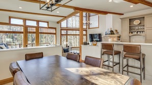 Uma sala de jantar espaçosa com uma grande mesa de madeira e cadeiras de couro, uma cozinha com banquetas e janelas amplas com vista para o exterior.