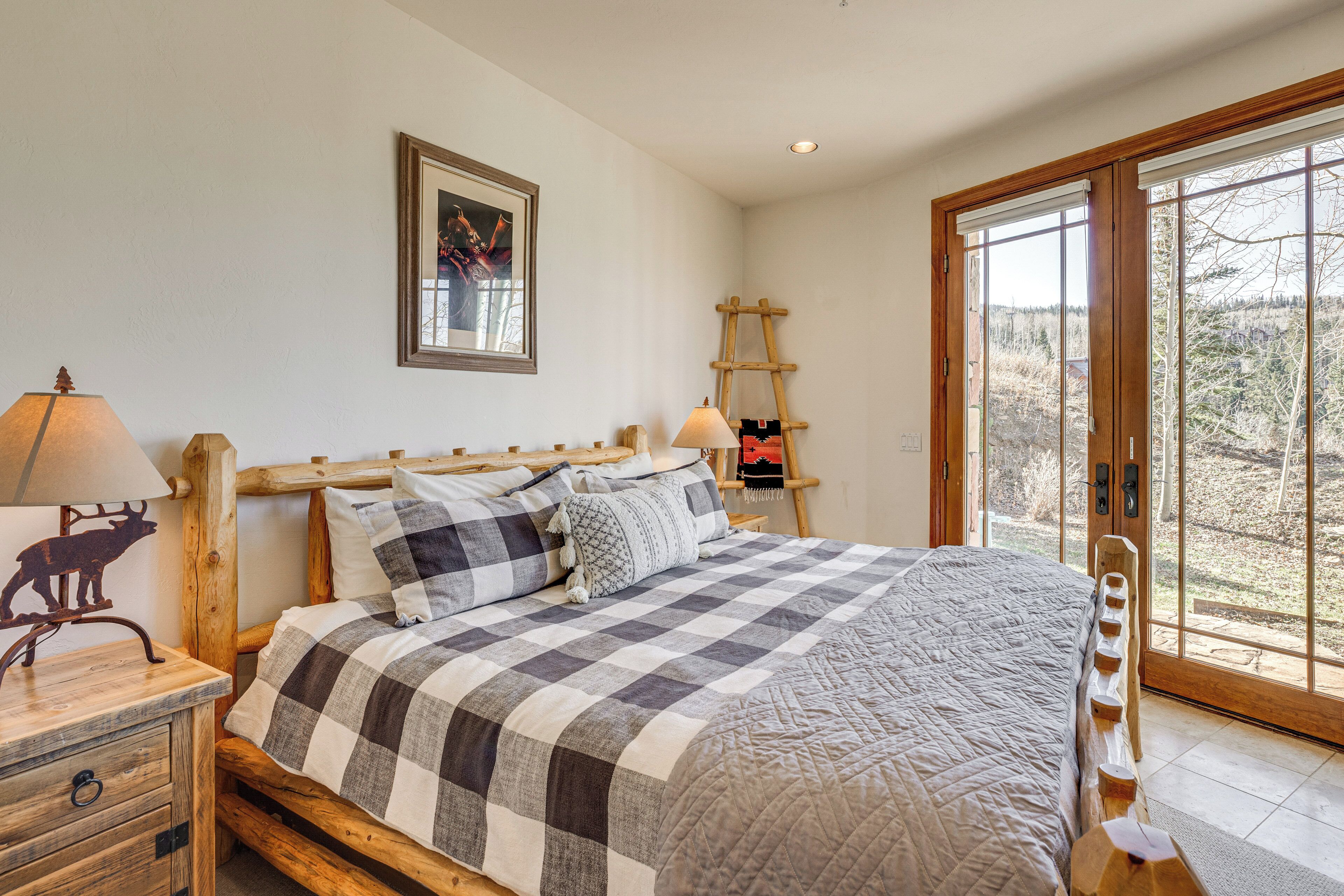 A bedroom with a wooden bed frame, checkered bedding, a nightstand with a lamp, and a ladder leading to a sliding glass door.
