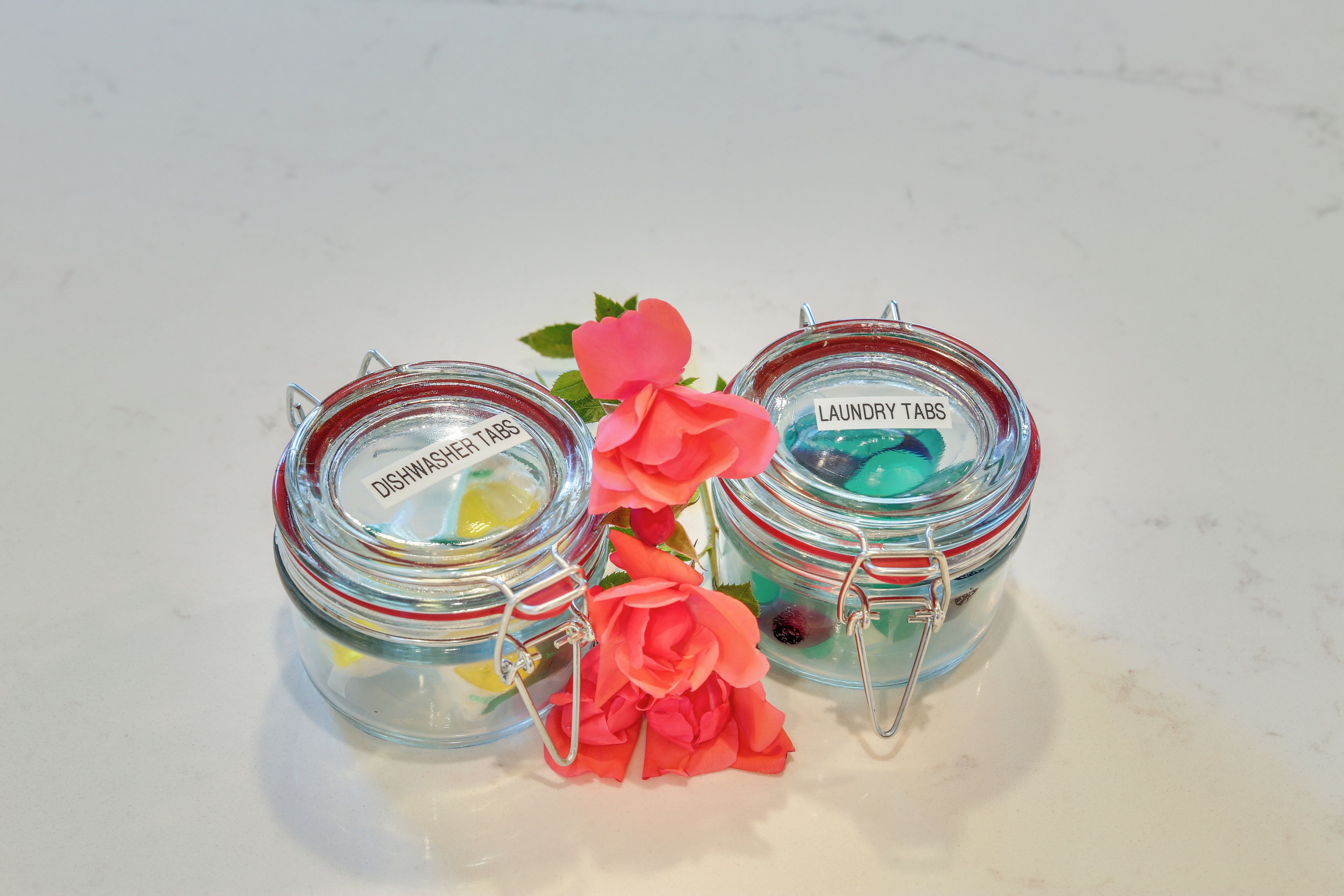 Tres pots de vidre amb tancaments metàl·lics, etiquetats com a “Pastilles per Rentavaixelles” i “Pastilles per Rentar Roba,” que contenen diferents articles.