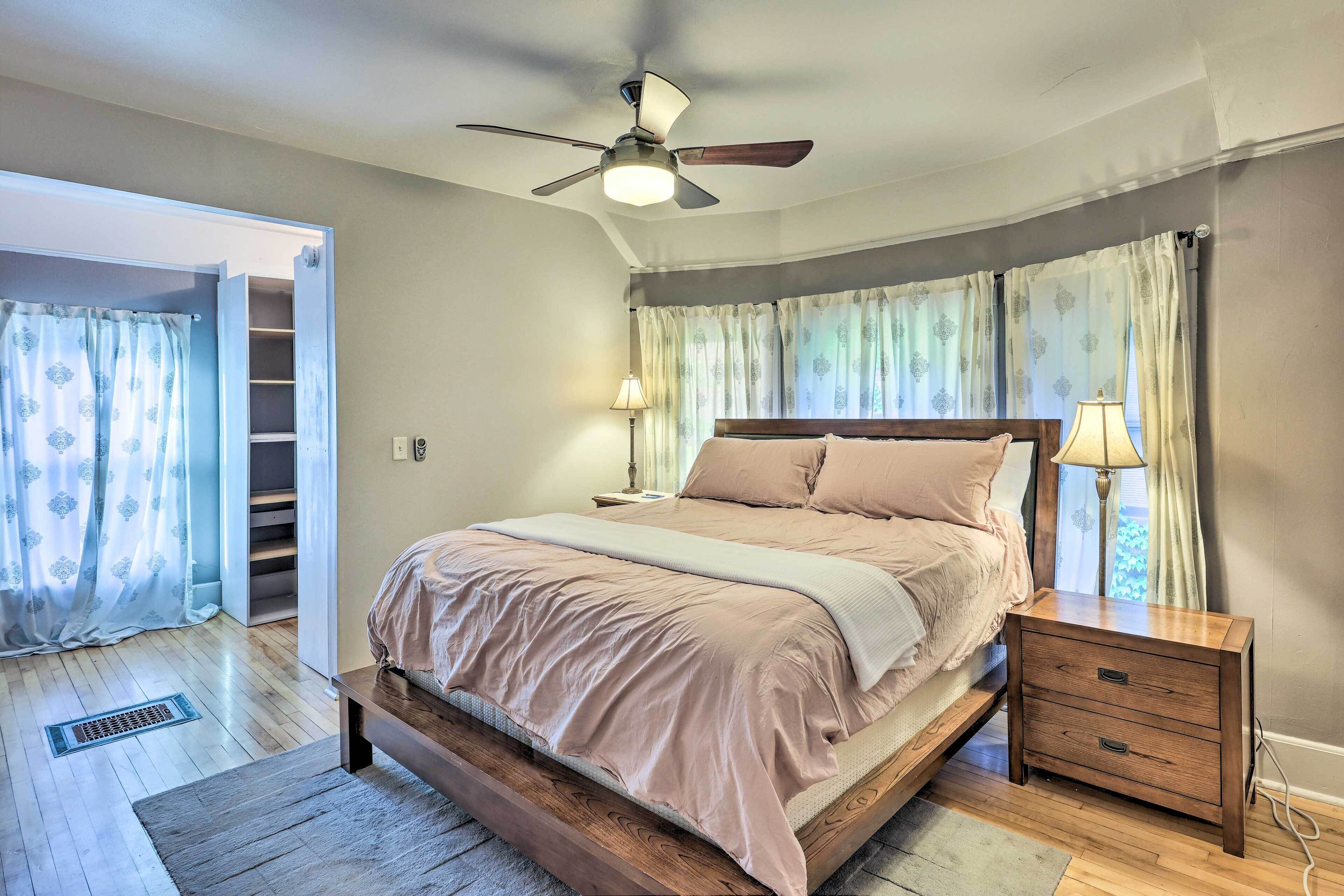 Quarto com cama, mesa de cabeceira, ventoinha no teto e janela com cortinas.
