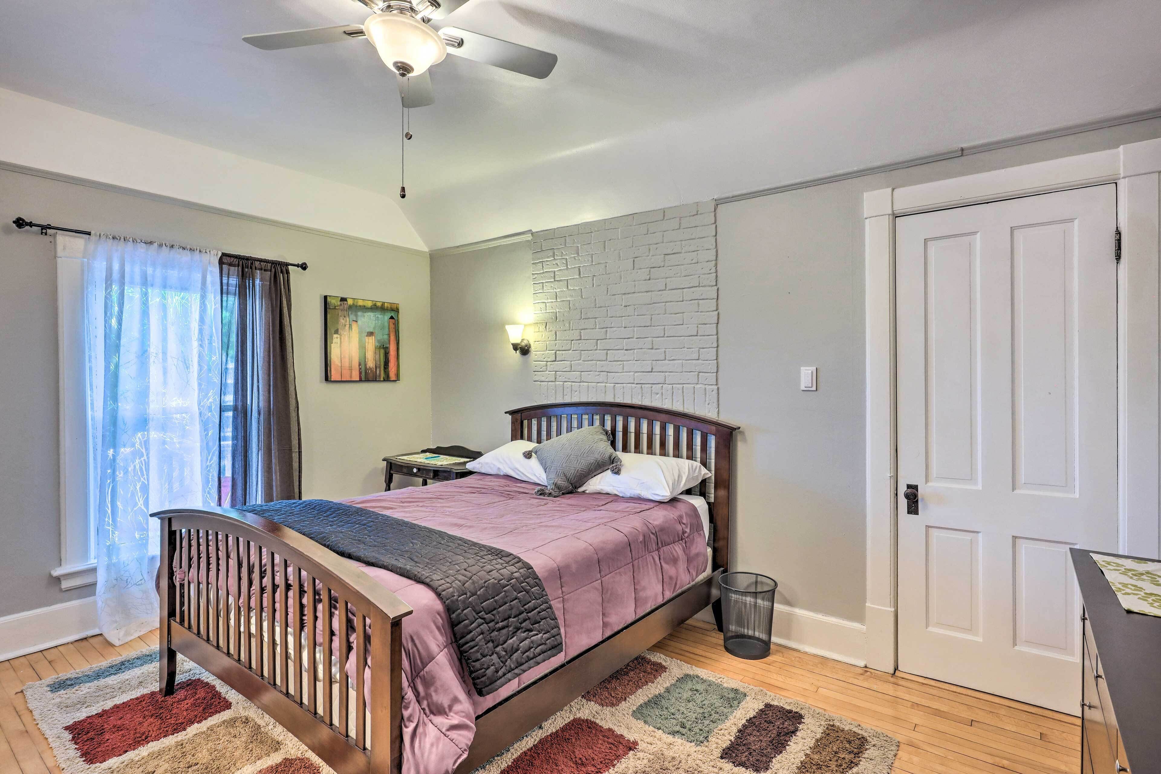 A bedroom with a bed, a ceiling fan, a nightstand, a wall-mounted lamp, a door, and a window with curtains.