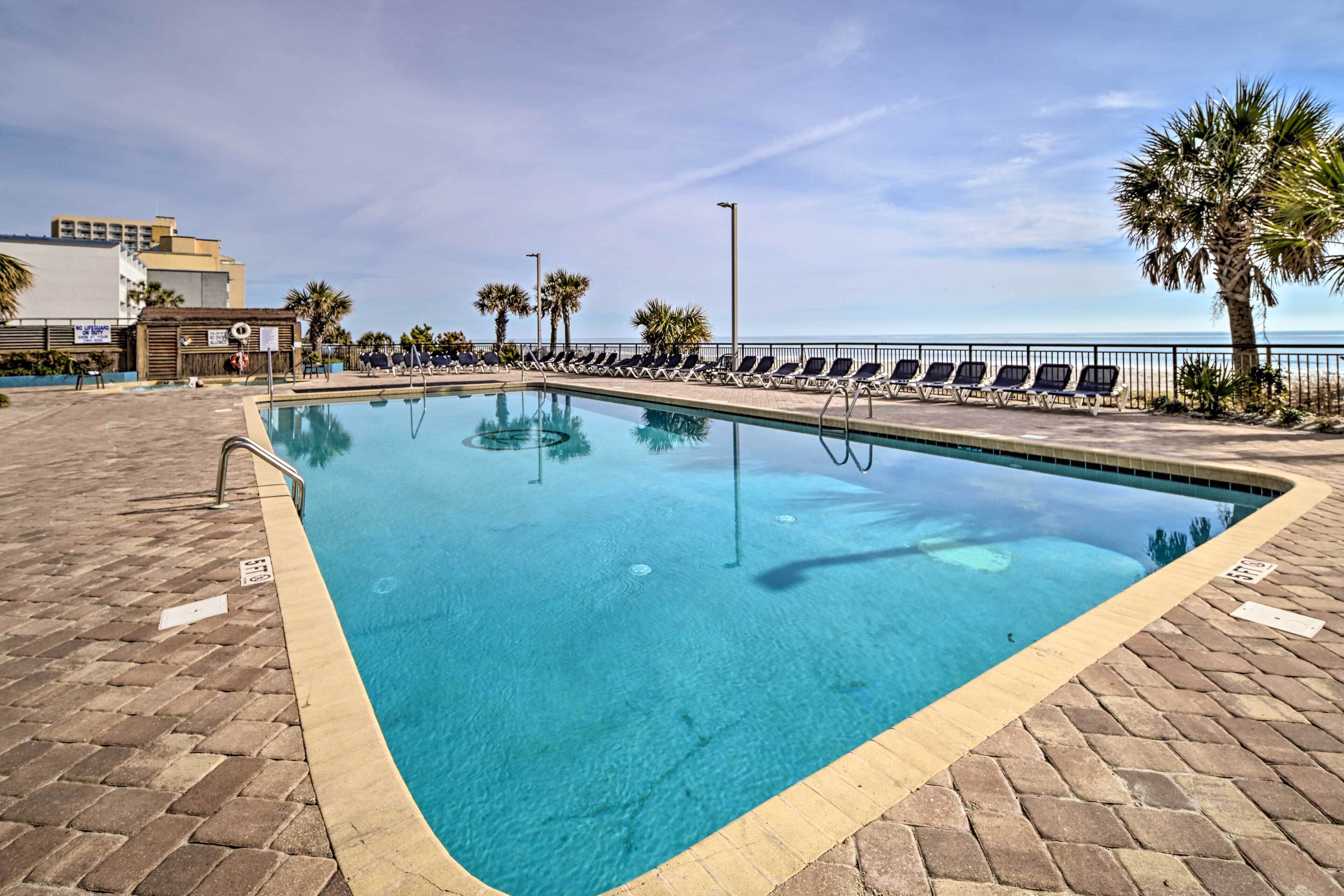 Une piscine rectangulaire à l’eau d’un bleu cristallin, entourée d’une terrasse en briques et bordée d’une rangée de chaises longues orientées vers l’océan.