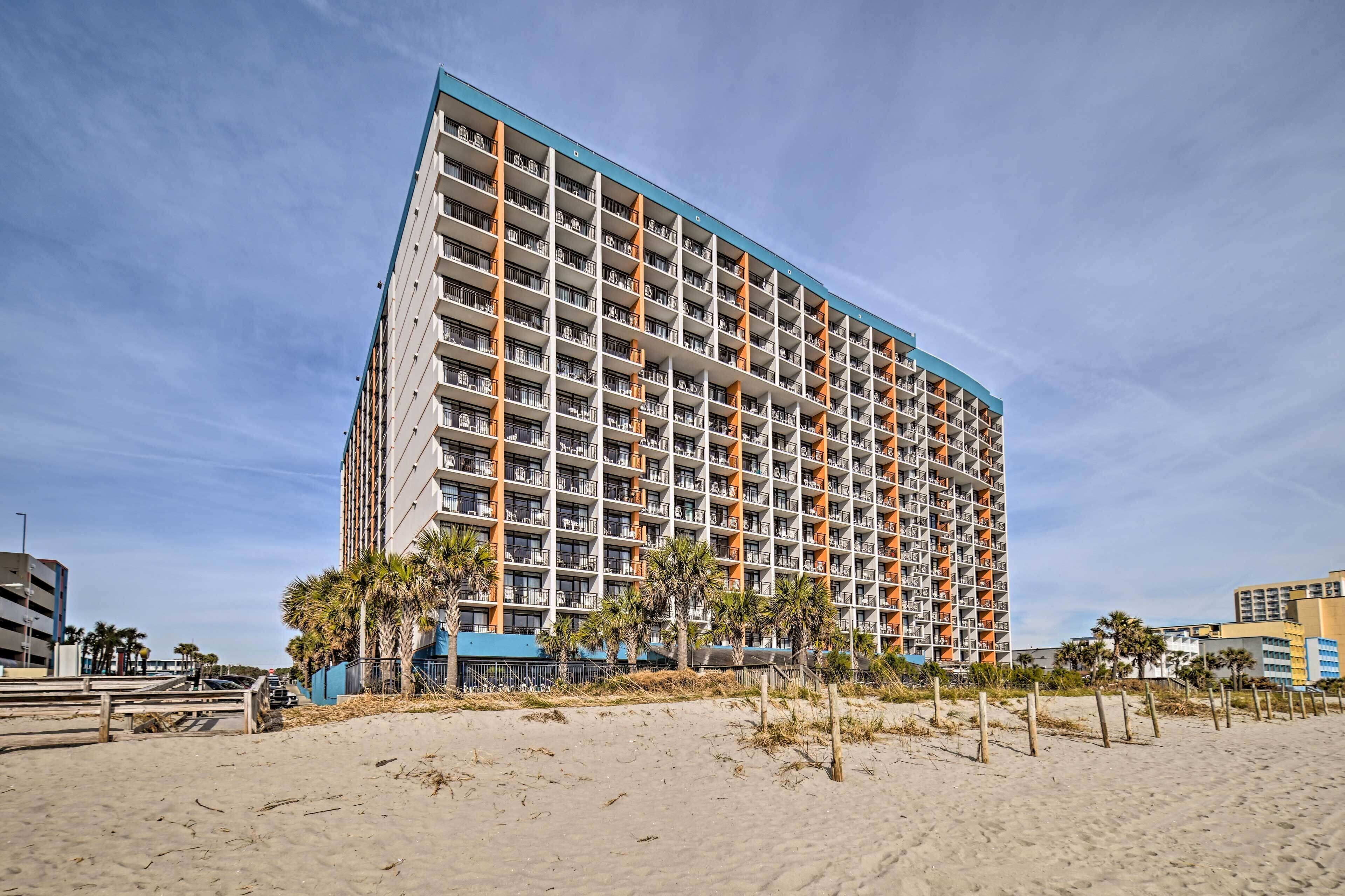 Un hôtel de grande hauteur doté de nombreux balcons, situé près d’une plage de sable bordée de palmiers.