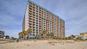 Un hôtel de grande hauteur doté de nombreux balcons, situé près d’une plage de sable bordée de palmiers.