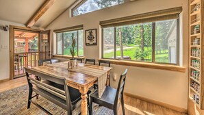 Un área de comedor con una mesa y sillas de madera, ventanas amplias con vistas a un paisaje verde y una estantería con frascos.