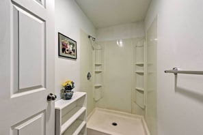 Une petite salle de bain blanche, avec une douche, une porte dotée d’un judas et une étagère sur laquelle est posé un vase de fleurs.
