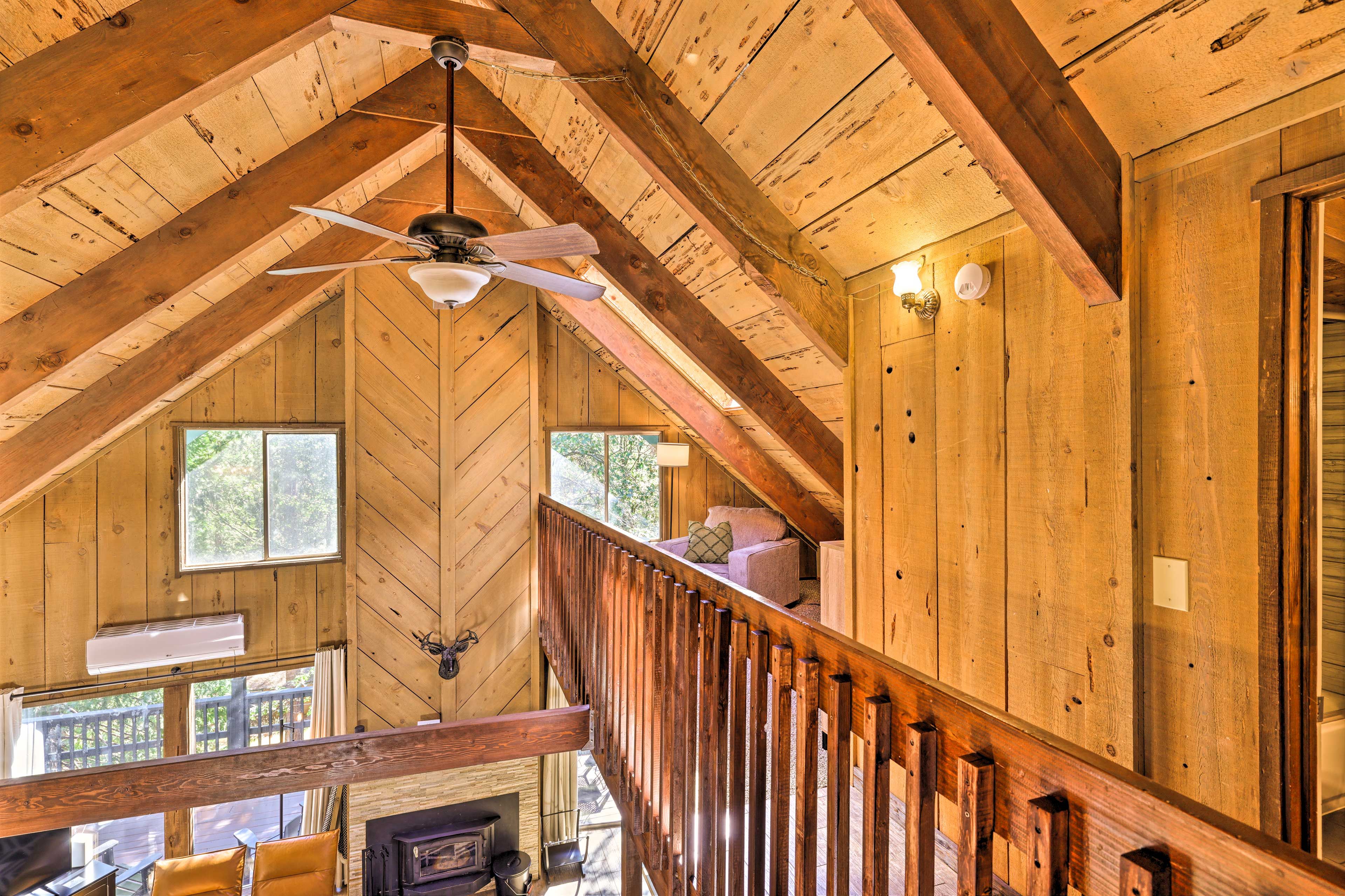 A wooden cabin interior with a loft bedroom, a fireplace, and a wooden staircase.