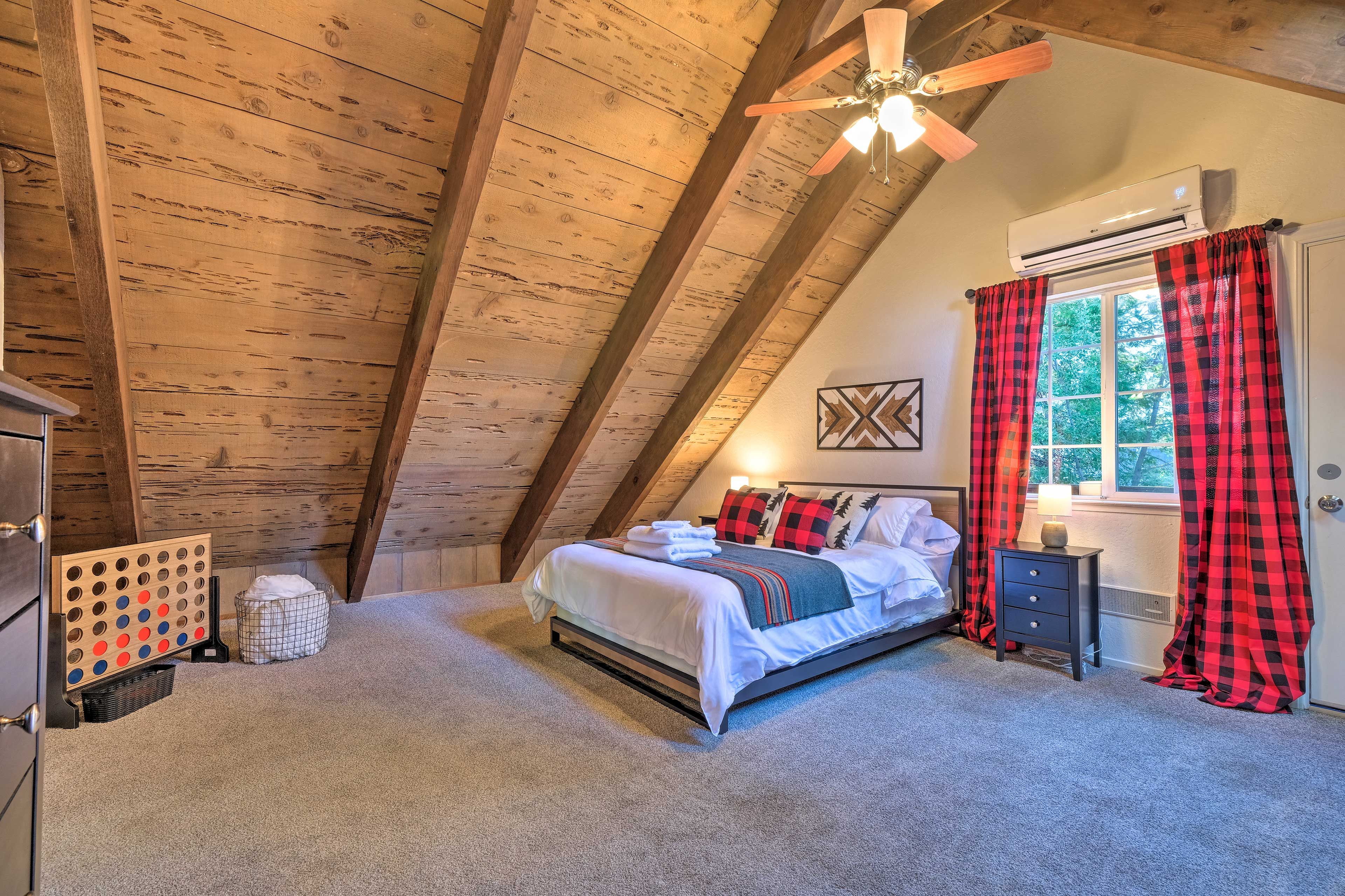 A bedroom with a sloped ceiling, a bed with white bedding, a nightstand, a window with red and black curtains, and a ceiling fan.
