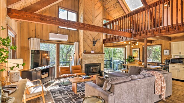 A spacious living room with a fireplace, wooden beams, and a large window overlooking a deck. - Near Trails: Cabin w/ 2-level Deck in Twain Harte (Twain Harte)