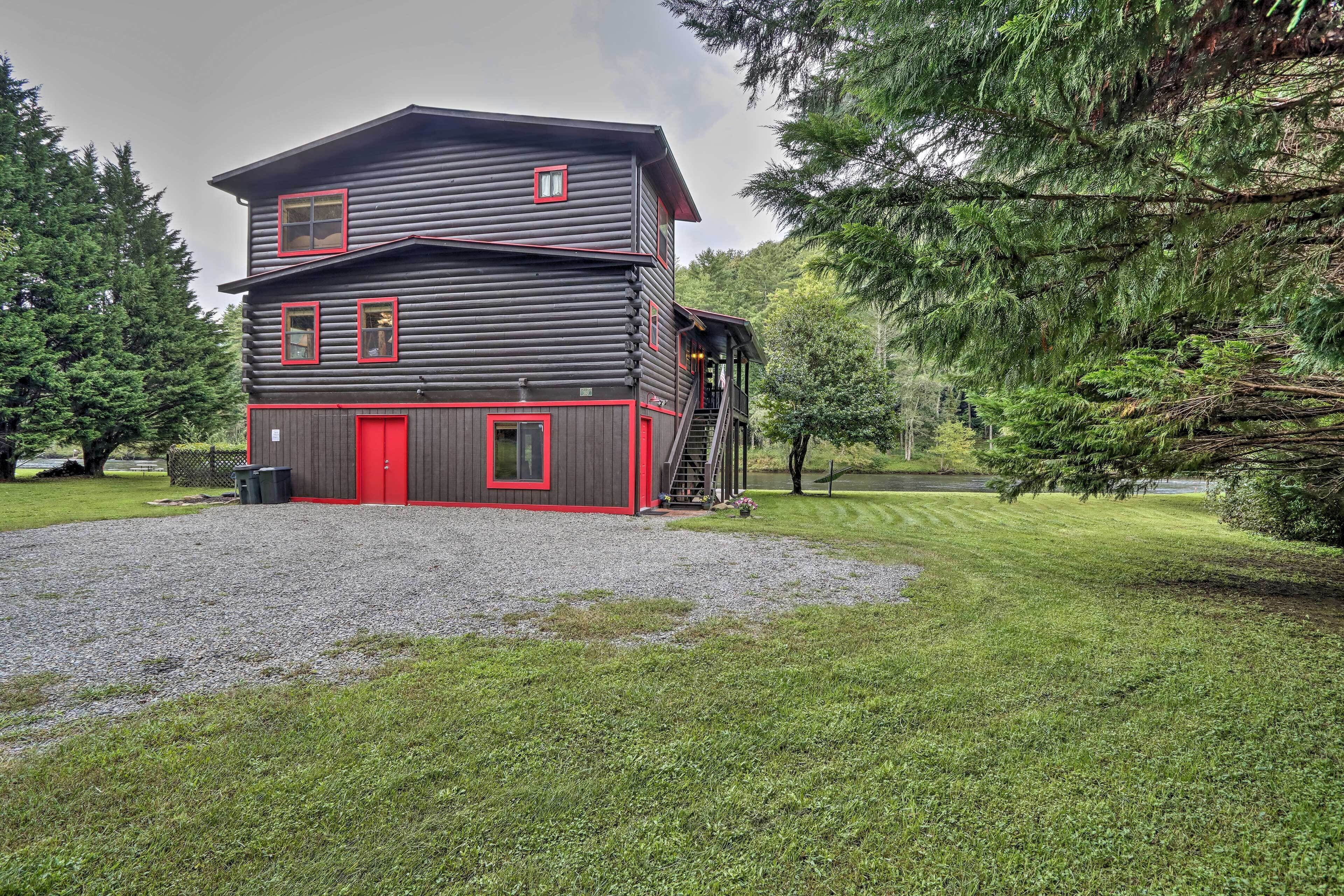 A two-story log cabin with a red door and windows, surrounded by a well-maintained lawn and dense forest.