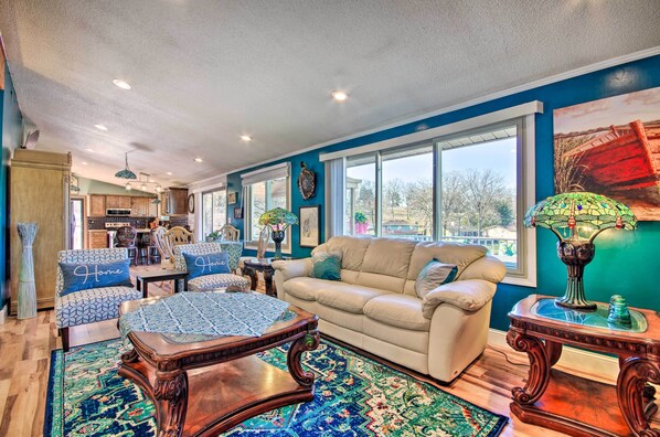 A spacious living room with a beige sofa, patterned armchairs, and a coffee table. A large window offers a view of the outdoors.