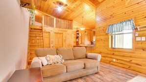 Ein gemütliches Blockhaushaus-Interieur mit Holzboden, einer Couch, einem kleinen Tisch und einem Fenster mit karierter Gardine.