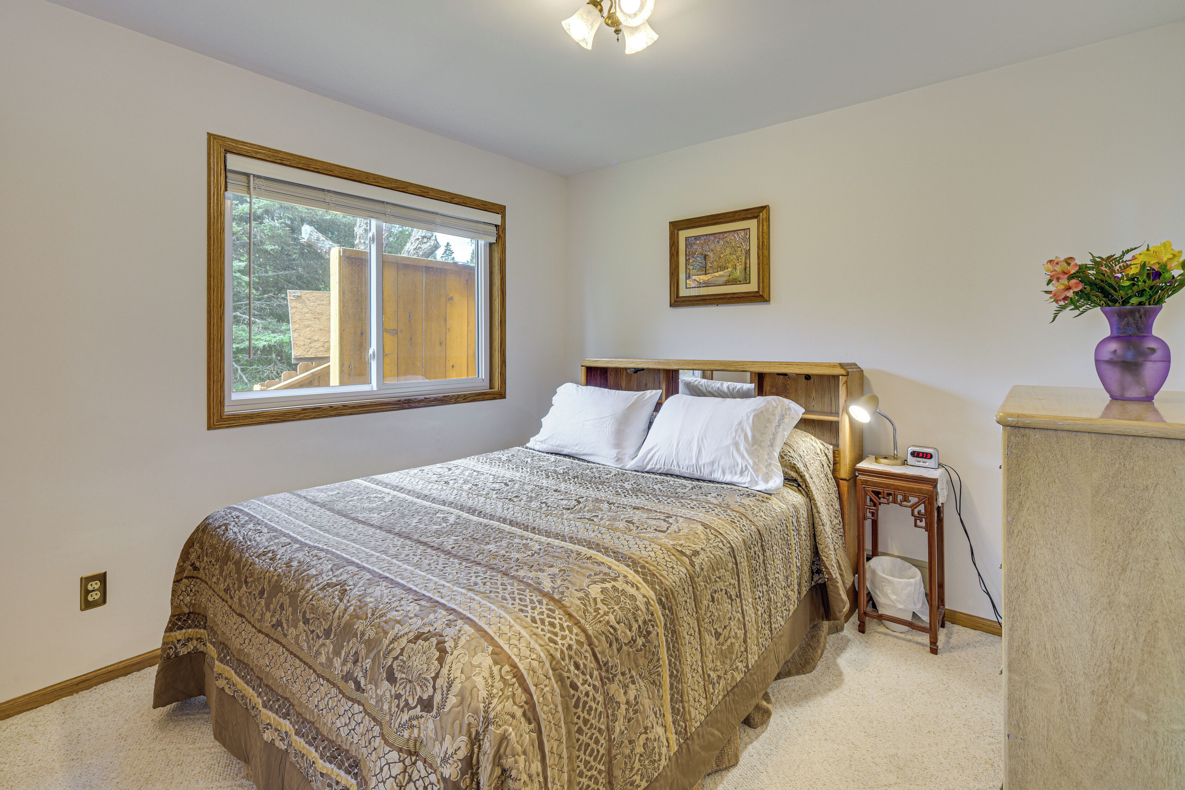 A bedroom with a large bed, a bedside table with a lamp, a framed picture on the wall, and a window with a view of trees.