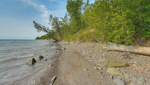 A pebbled beach with a fallen tree trunk, rocky shoreline, and dense greenery. - 15 Mi to Duluth: Lake Superior Beachfront Home! (Duluth)