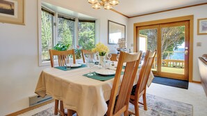 A dining area with a table set for four, wooden chairs, a chandelier, and large windows with a view of greenery and a wooden deck outside. - 15 Mi to Duluth: Lake Superior Beachfront Home! (Duluth)