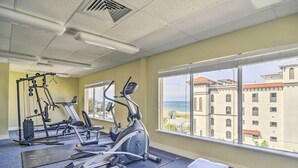 A gym with various exercise equipment, a large window with a view of buildings and the sea.