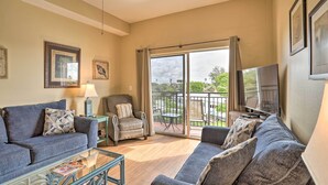 A spacious living room with a sofa, armchair, and a glass coffee table. A television is mounted on the wall, and there is a balcony with a table and chairs outside.