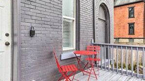 Un balcon avec un mur en briques grises, une fenêtre, ainsi qu’une table et une chaise en métal rouge.