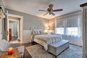 Une chambre à coucher comprenant un lit, un ventilateur de plafond, une table de chevet avec une lampe, une fenêtre avec des stores et une porte donnant sur une autre pièce.