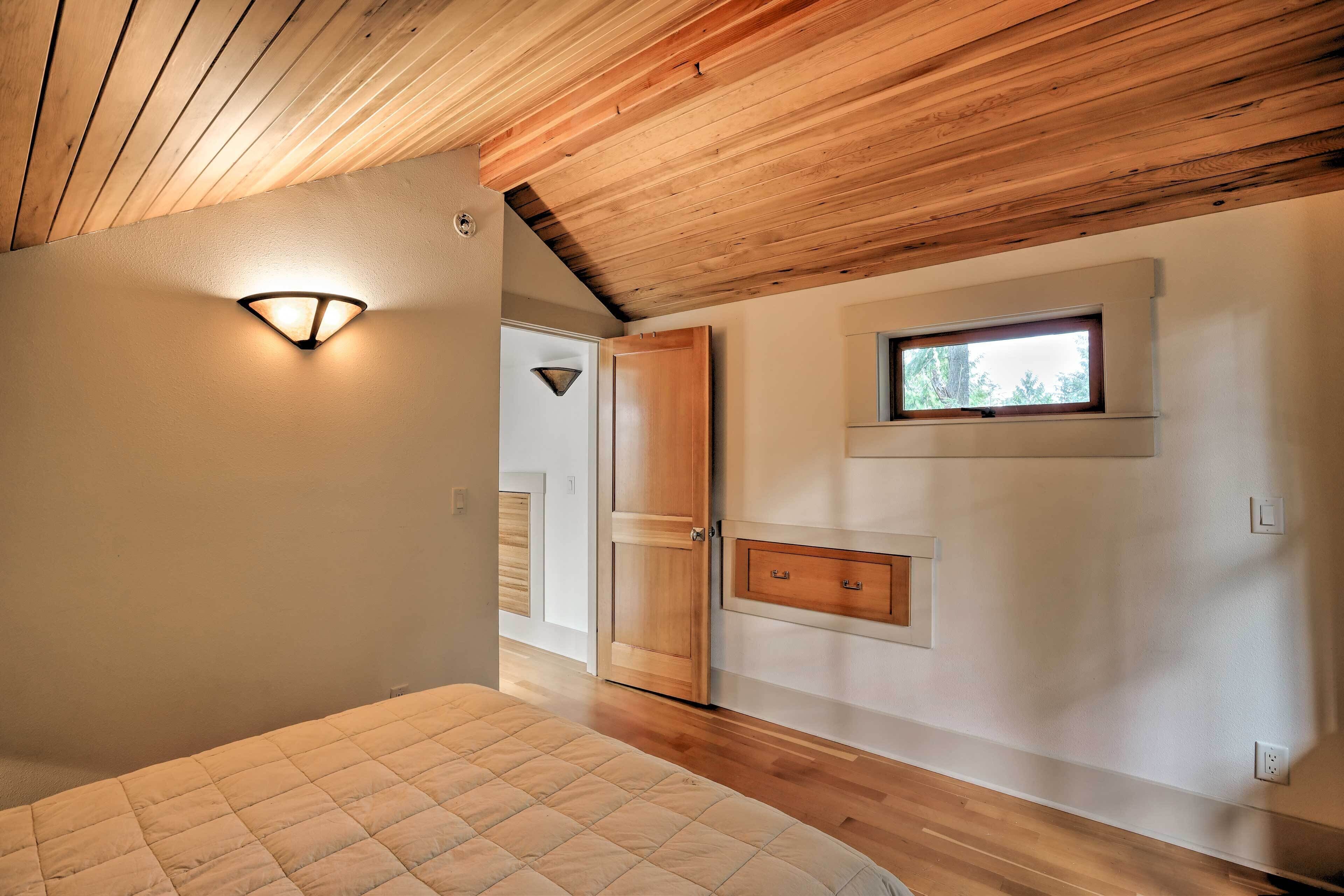 A bedroom with a wooden ceiling, a bed with a quilted cover, a wall-mounted light fixture, a window, and a door.