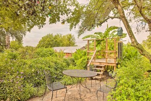 Een patio in de tuin met een metalen tafel en stoelen, omgeven door weelderige begroeiing en een houten constructie.