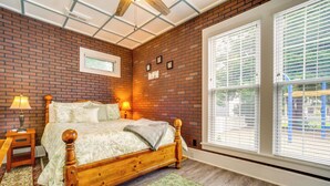Ein Schlafzimmer mit Backsteinwand, einem hölzernen Bett, zwei Nachttischlampen, einem Fenster mit Jalousien und einem kleinen Bild an der Wand.