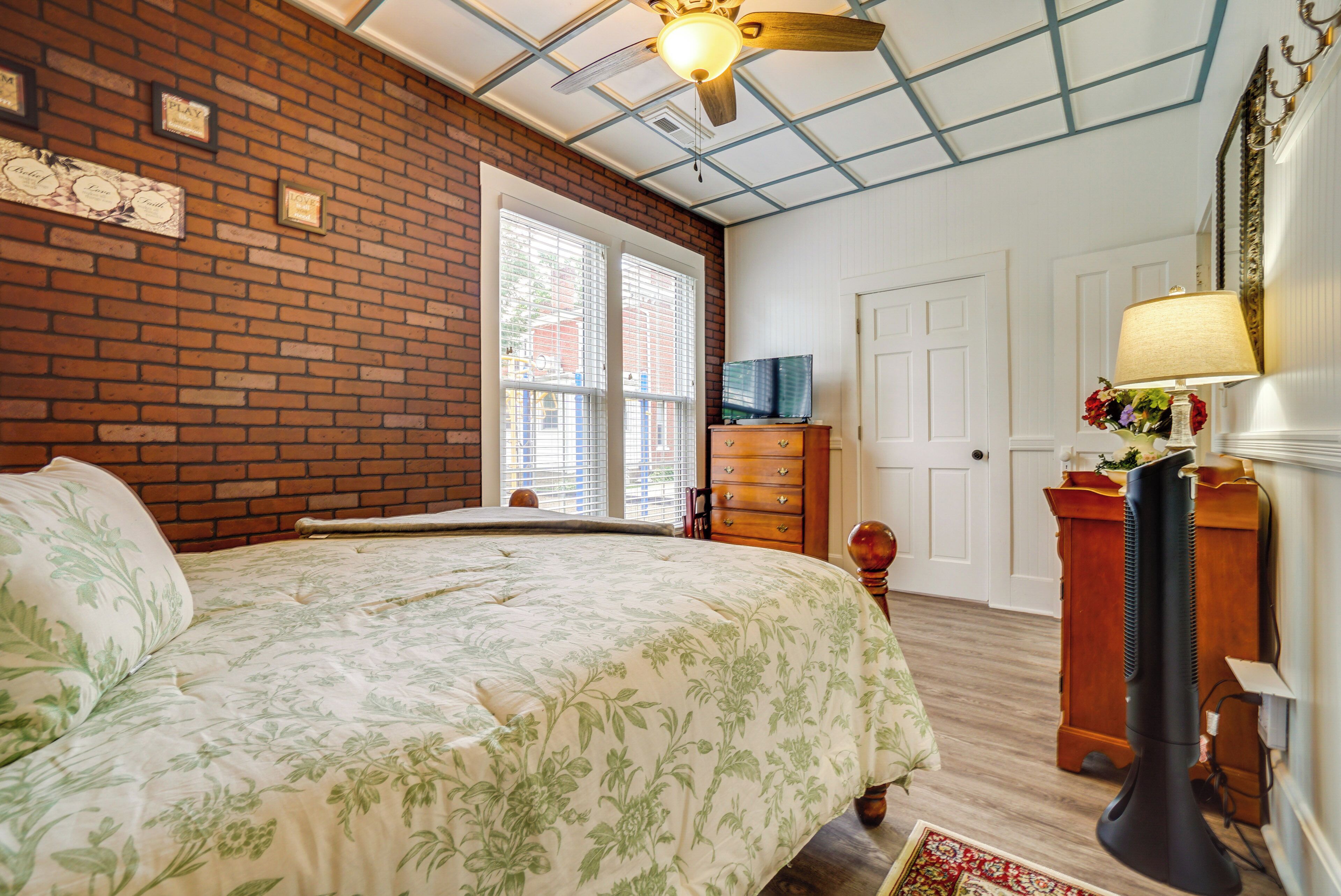 A bedroom with a brick wall, a wooden bed, a ceiling fan, a dresser, and a television.