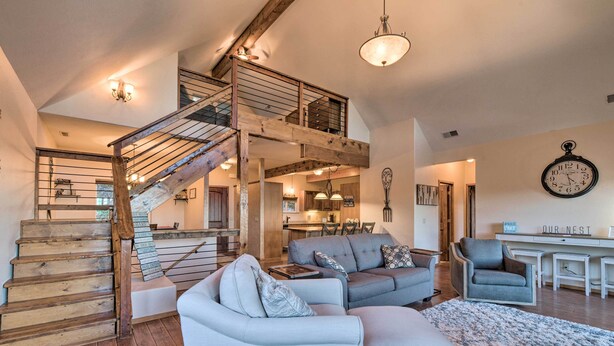 A spacious living area with a wooden staircase, a grey sofa set, and a dining area with a clock on the wall.