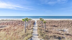 Uma trilha de areia leva a uma praia com palmeiras e um céu azul límpido.