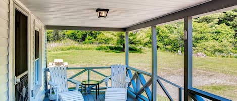 A covered porch with a table and four Adirondack chairs, overlooking a grassy area.