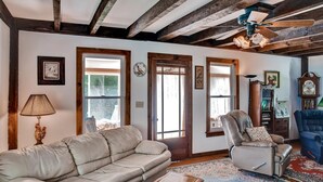 Ein Wohnzimmer mit einer Couch, einem Sessel und einem Couchtisch. Zwei Fenster mit Vorhängen, ein Deckenventilator und eine Uhr an der Wand sind ebenfalls zu sehen.