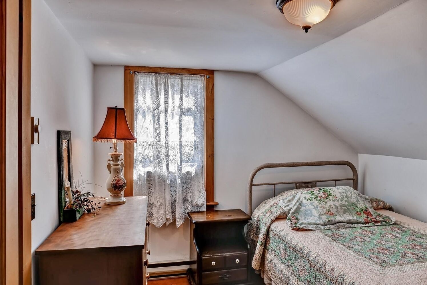 Un dormitorio pequeño y acogedor con techo inclinado, una cama individual con ropa de cama floral, una mesita de noche con una lámpara y una ventana con cortinas de encaje.
