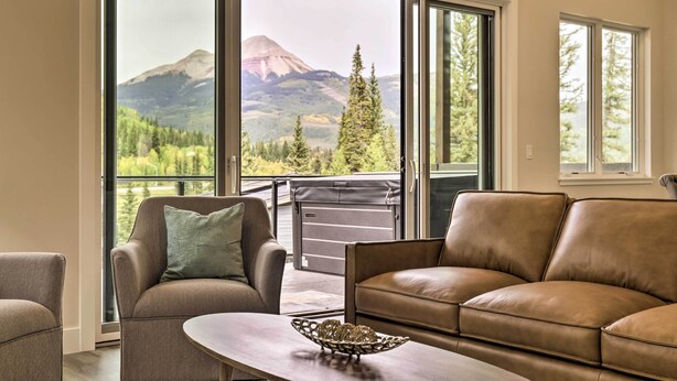 A living room with a brown leather sofa, a matching armchair, and a coffee table. Large sliding glass doors lead to a balcony with a mountain view.