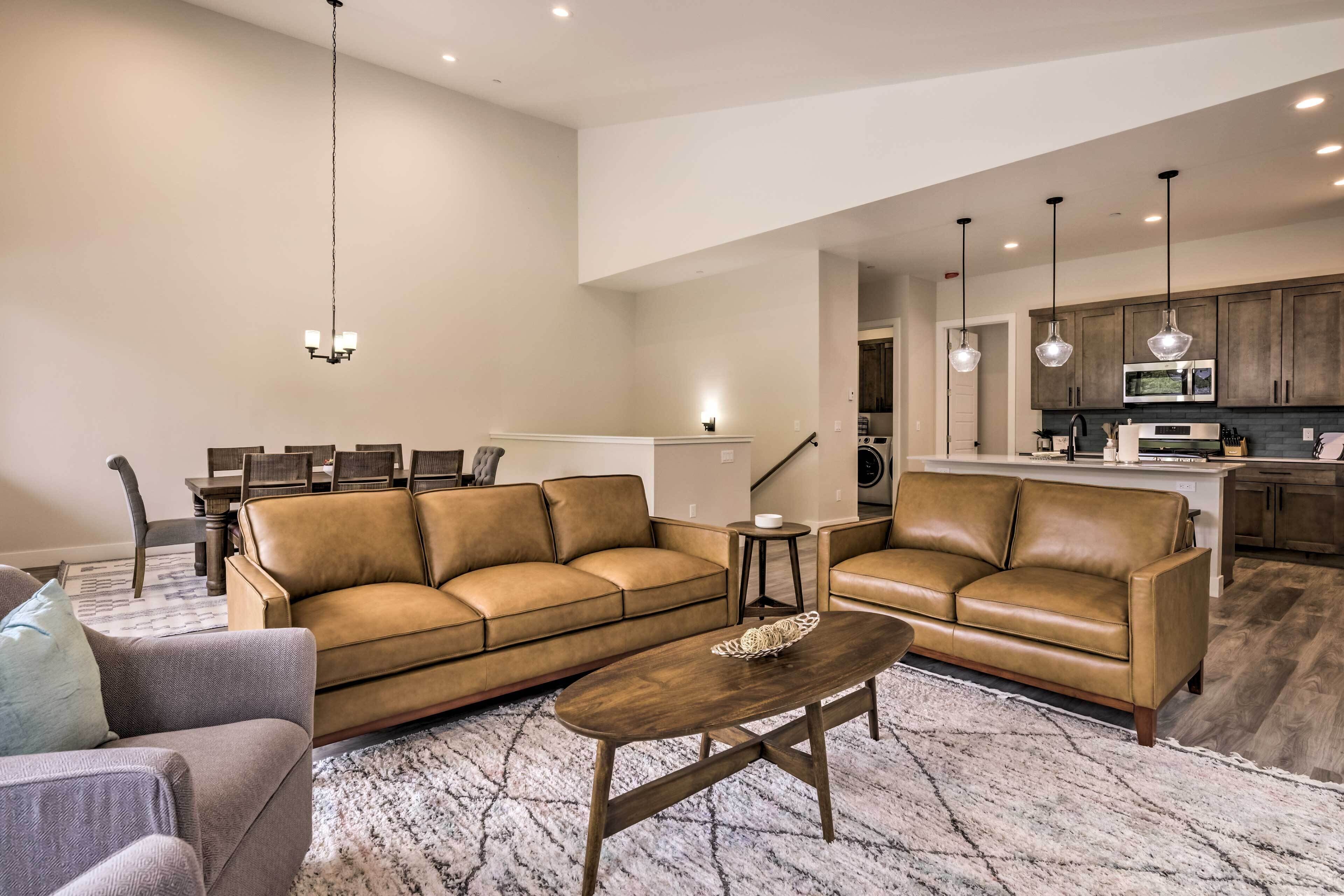 Ein geräumiges Wohnzimmer mit einem braunen Ledersofa, einem Holztisch und einem grauen Sessel. Im Hintergrund ist ein Essbereich mit Tisch und Stühlen zu sehen.
