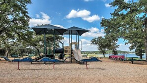 A playground with slides and swings, surrounded by trees and a body of water. - Beach & Pool Access at Lake Travis Family Getaway! (Leander)