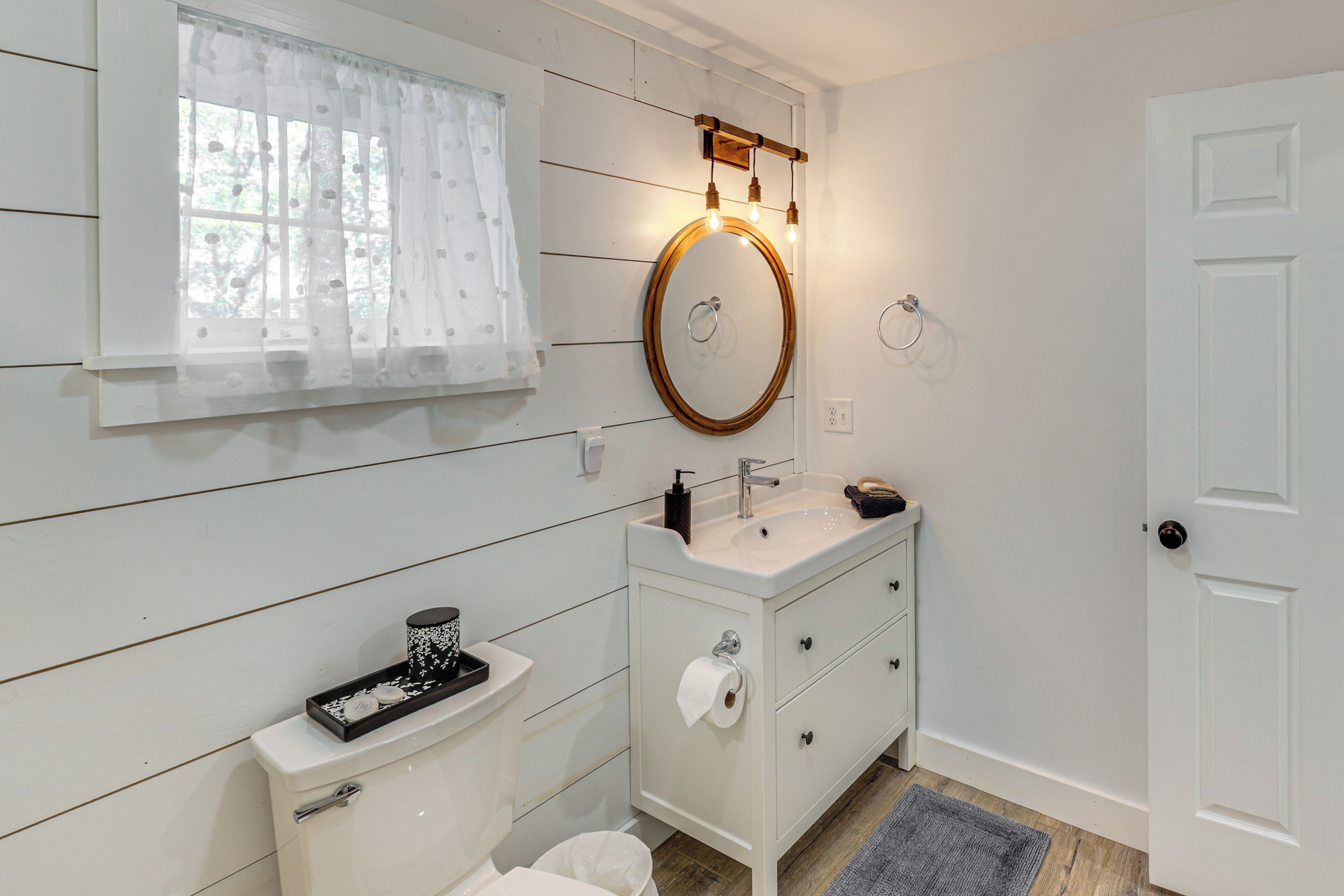 A bathroom with a white vanity, a round mirror with a wooden frame, a toilet, and a window with sheer curtains.