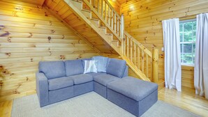 Un intérieur en bois avec un escalier, un canapé d’angle gris et une fenêtre avec des rideaux.
