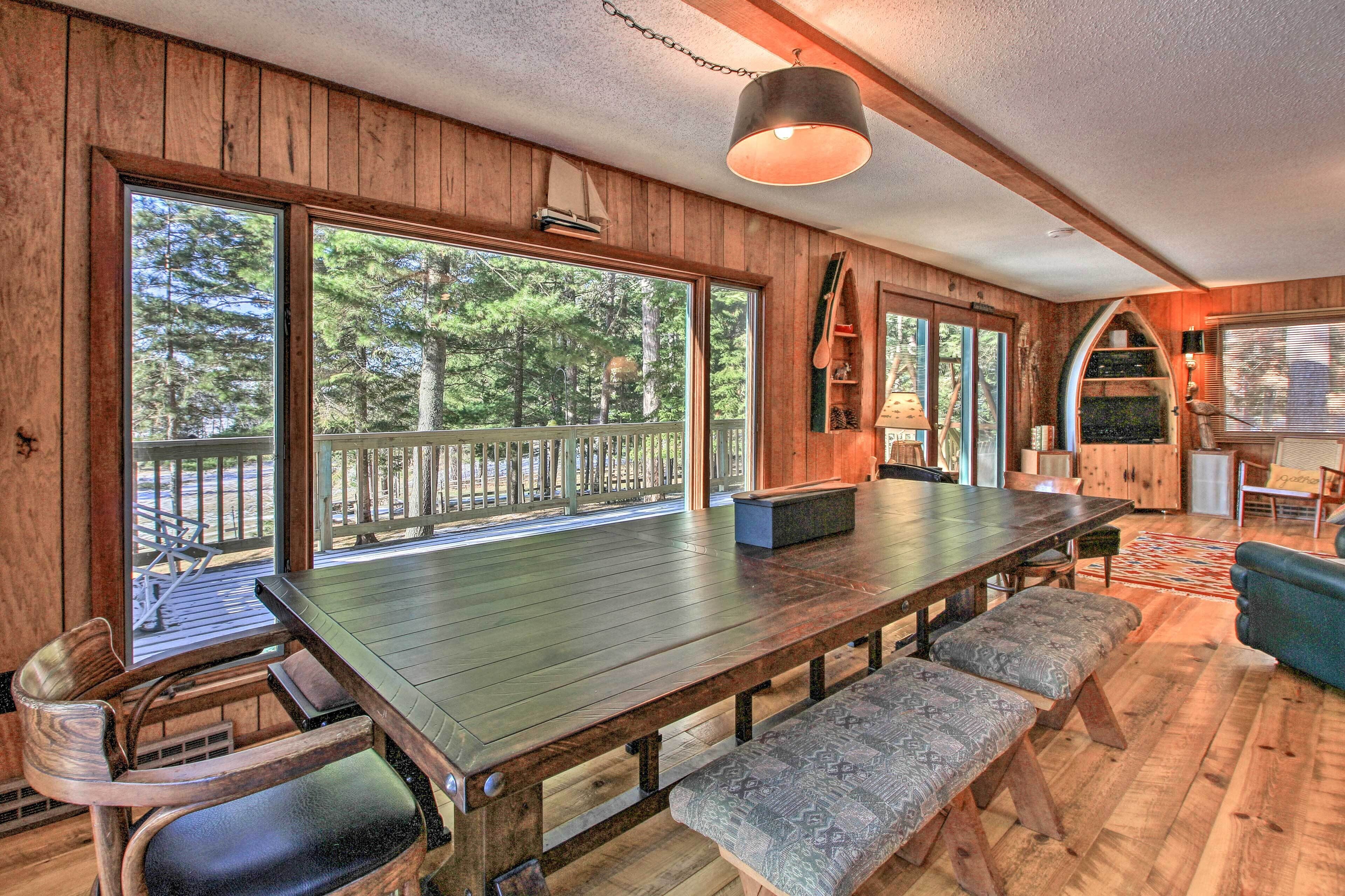 A wooden interior with a large dining table, benches, and a view of the forest through large windows.