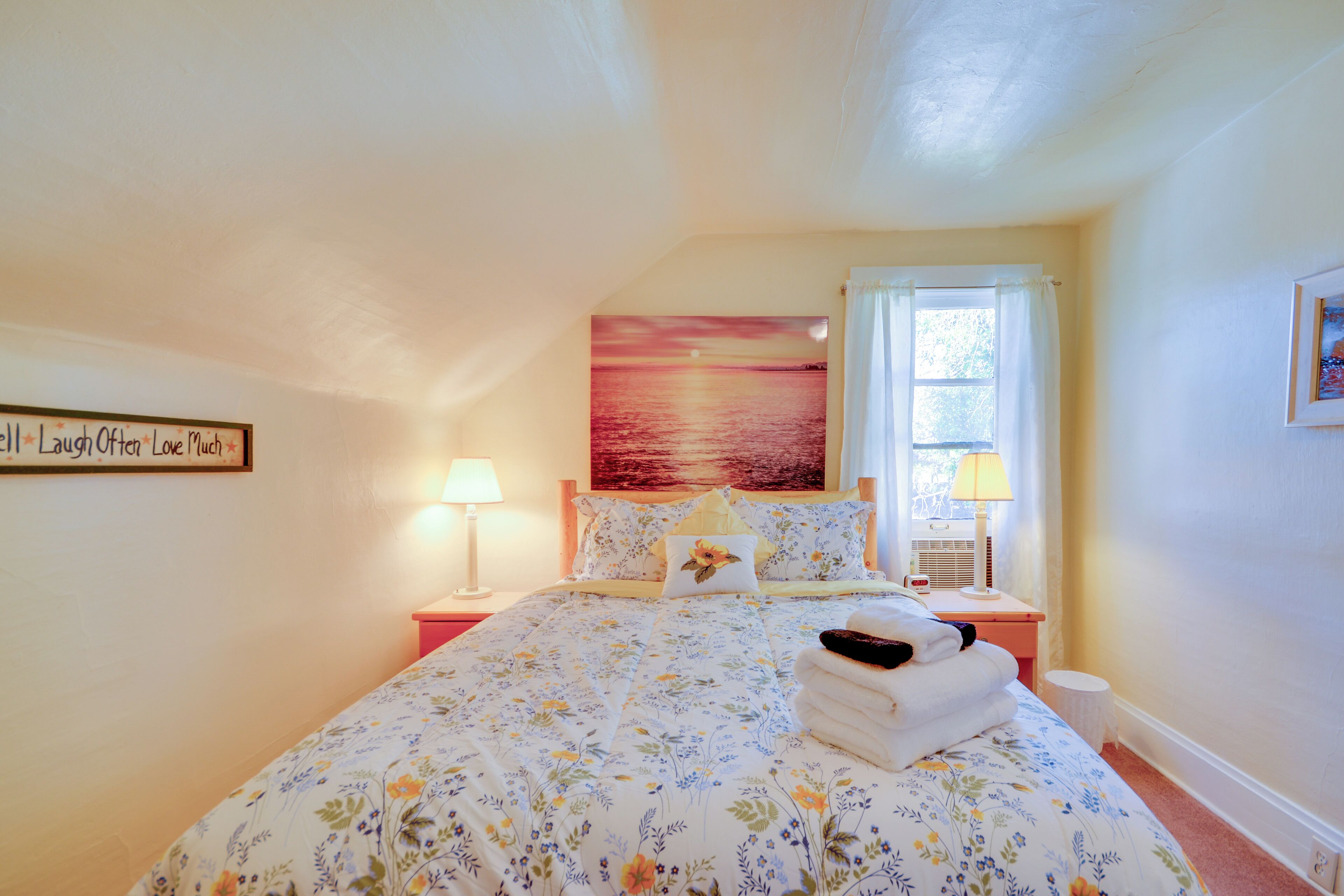 Een slaapkamer met een bloemenbedekker, een schilderij van een zonsondergang, een bedlampje en een raam met gordijnen.