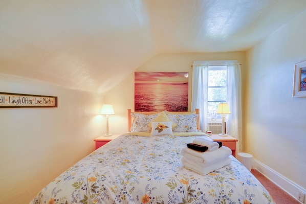 A bedroom with a floral bedspread, a painting of a sunset, a bedside lamp, and a window with curtains.