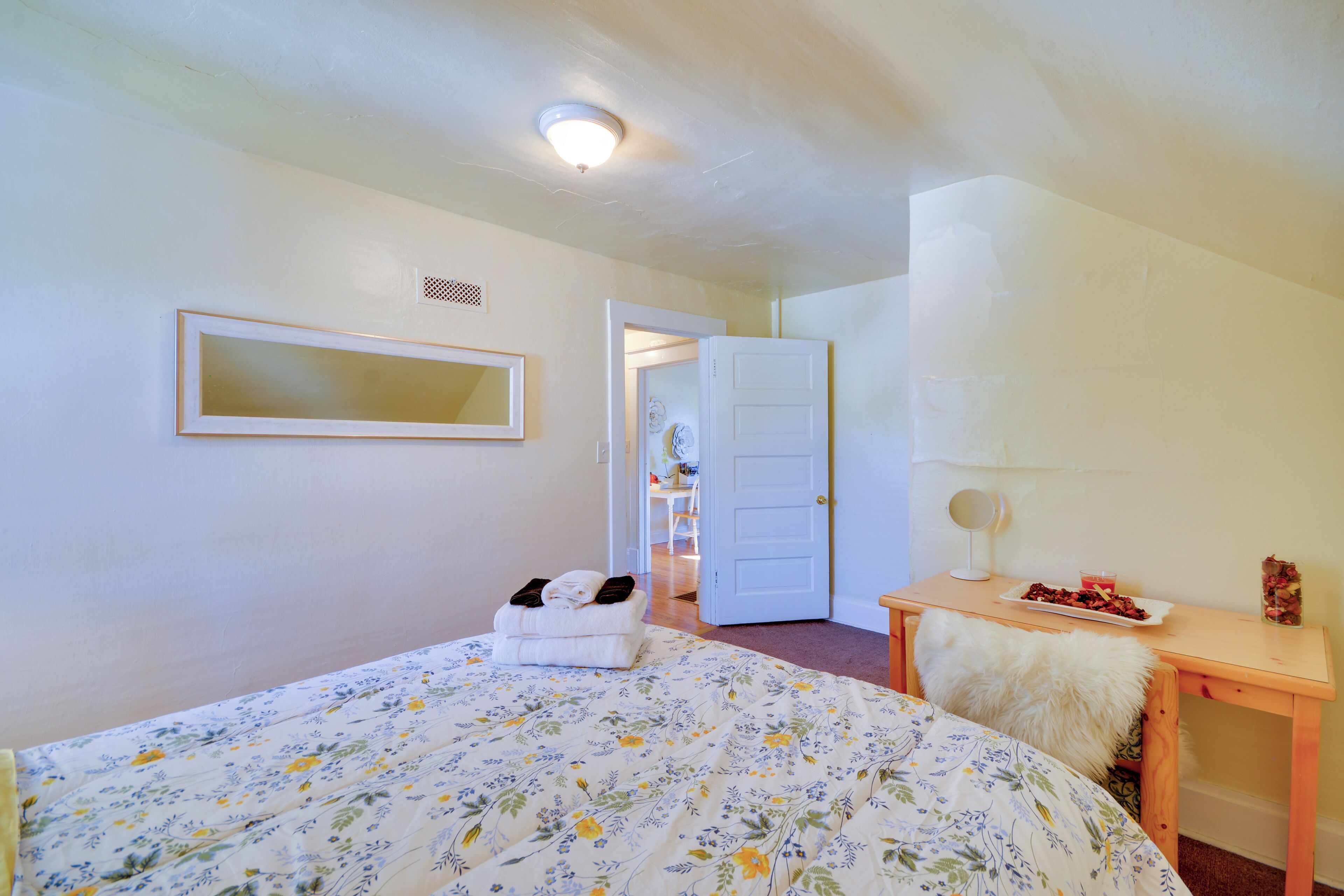 Un dormitorio con una cama, una mesita de noche con lámpara, un espejo y una puerta que da a otra habitación.