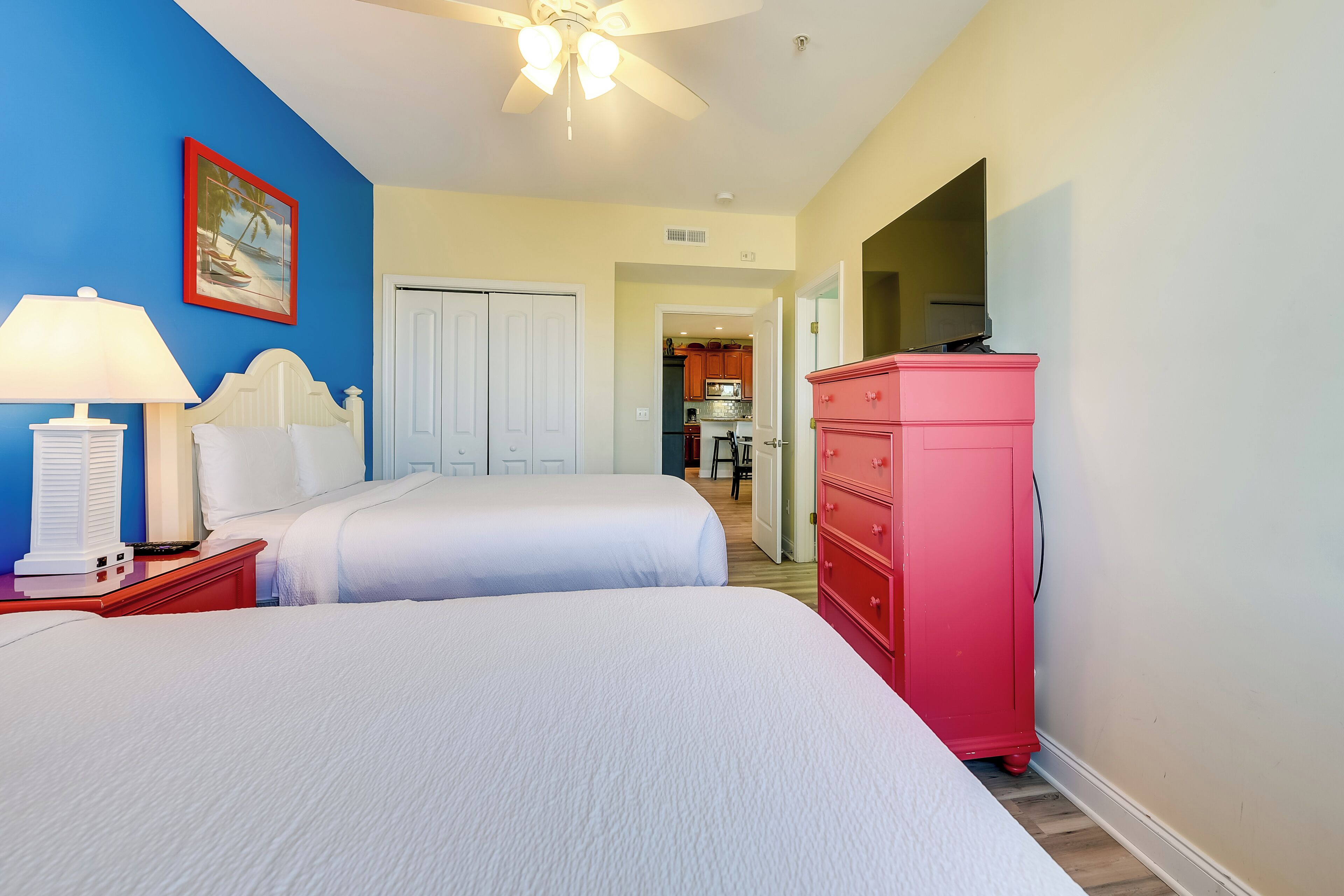 A hotel room with two beds, a red dresser, a ceiling fan, and a painting on the wall.