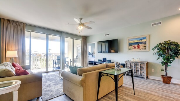 A spacious living room with a sofa, a coffee table, and a TV. There is a balcony with a view of the city. - Walk to Shore: Roomy Condo in North Myrtle Beach! (North Myrtle Beach)