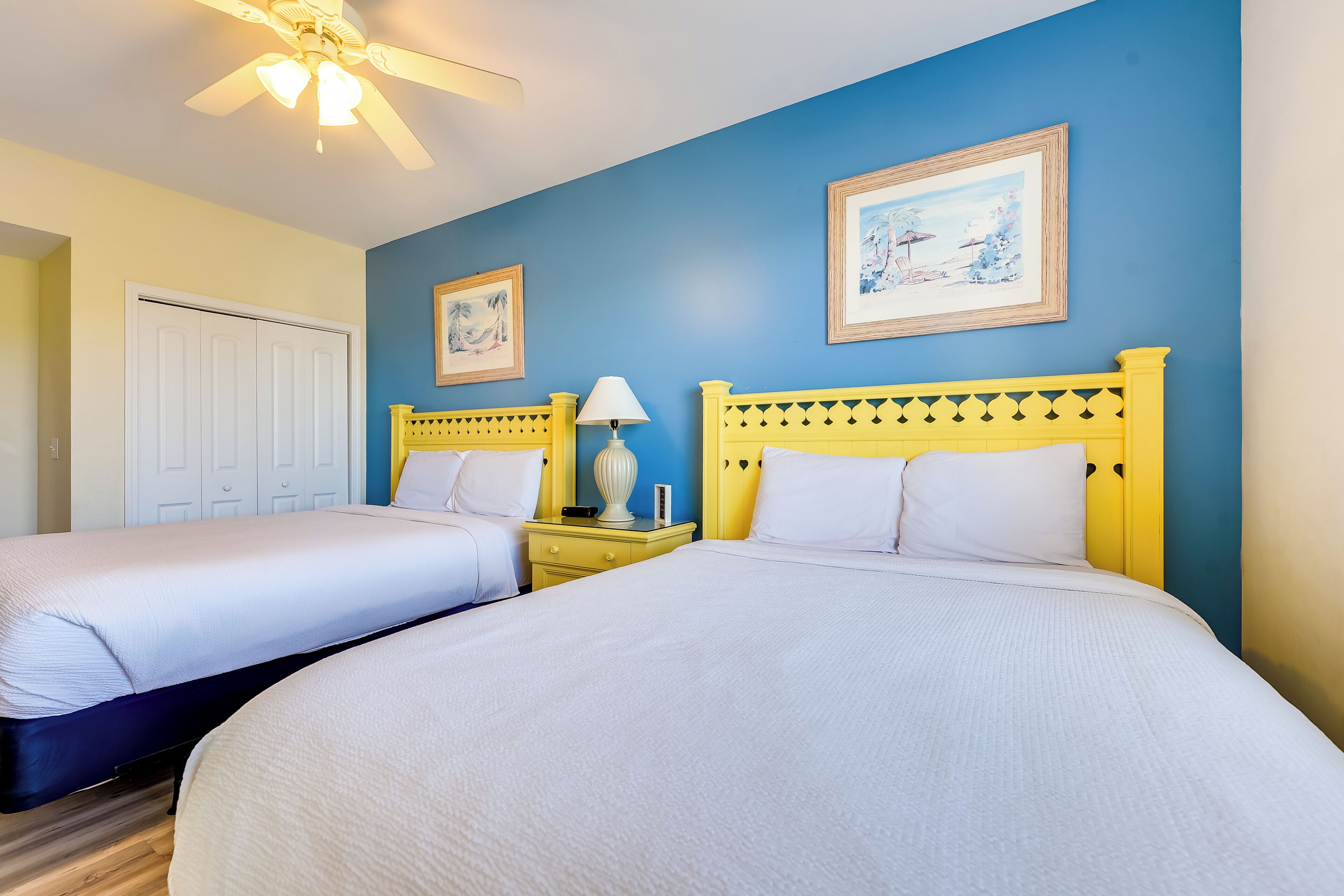 A hotel room with two beds, a ceiling fan, a nightstand with a lamp, and framed artwork on the walls.